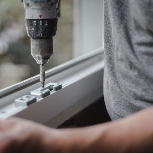 Young caucasian man installs metal fittings for plastic window frame with a drill, tightening a screw in a room where renovations are taking place, close-up side view with selective focus. The concept of home renovation, washing window frames.